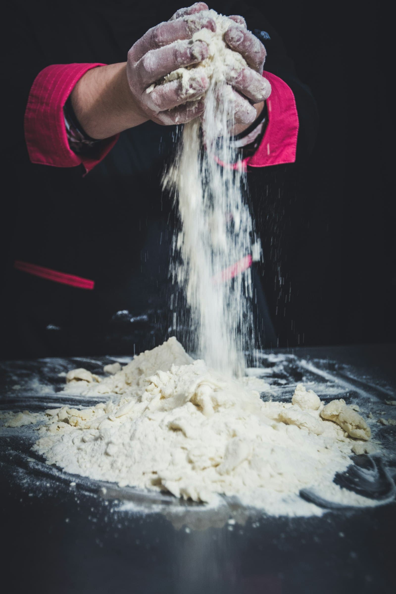 Flour-dusted hands letting a cascade of flour fall dramatically over dough on a dark surface
